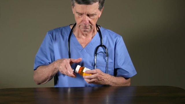 Senior doctor in blue scrubs with stethoscope pouring white tablets of oxycodone from prescription bottle onto desk to illustrate opioid addiction