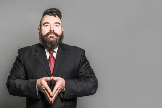 Adult Bearded Man In A Suit With Folded Triangle Hands On A Gray Background