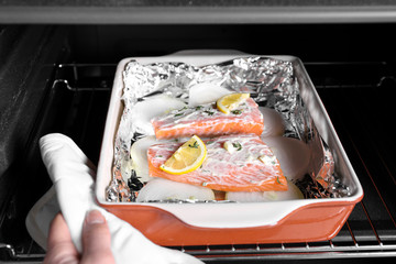 Woman putting baking dish with delicious salmon fillet into oven