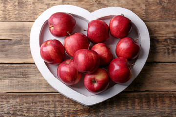 Plate in shape of heart with fresh red apples on wooden background