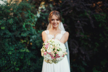 Bride reaches out her hands with pink wedding bouquet standing before green bushes