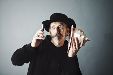 senior in a hat with shell listening noise of sea and phone