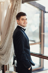 Smiling groom in black suit stands in the rays of morning sun before the window