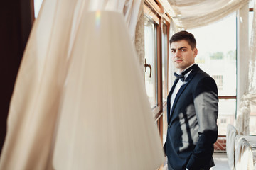 Look from behind the curtain at handsome groom standing before tall window