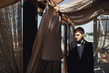 Thoughtful groom with black bow tie stands before panoramic window