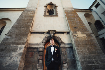 Tall groom in black suit stands before old wooden wall
