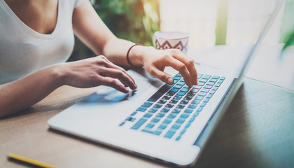 Young woman working at home on modern computer while sitting at the wooden table.Female hands typing on laptop keyboard.Concept of young modern people using mobile devices at home.Blurred background.