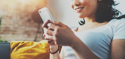 Closeup view of young smiling Asian woman sitting on sofa at home, wearing white tshirt and typing hands on smartphone.Horizontal blurred, flares effect.Wide.Selective focus.