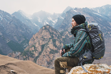 Tired bearded hipster with binoculars in his hands sits on a stone among the mountains and looks out into the distance