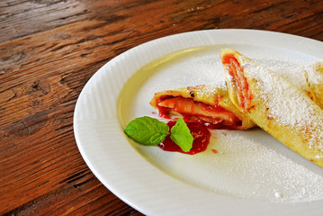 Rolled pancakes with powdered sugar and strawberry jam. French crepes on white plate on wooden background