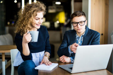 Fototapeta premium Young couple surfing the Internet on a laptop. Woman drink a cup of coffee and speak with man. Morning in modern office.