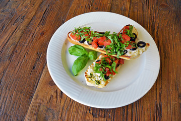 Preparing delicious Italian tomato bruschetta with chopped vegetables, olives, cheese and oil on grilled or toasted crusty baguette sprinkled with seasoning and spices on a plate on wooden background