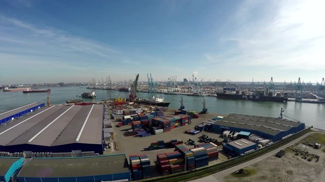 Aerial bird view flying over harbor area industrial zone with intermodal containers and in background showing cranes for loading and unloading containers then flying past boat with helicopter platform