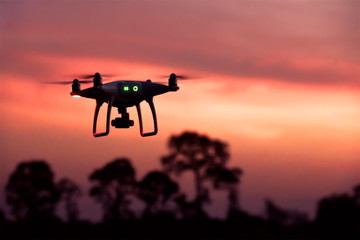 Silhouette of modern RC Drone / Quadcopter with camera flying above tree background at sunset