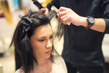 Fototapeta premium Young woman and hairdresser with hair iron making hairdo at hair salon