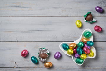 Chocolate Easter eggs wrapped in colorful foil in a butterfly shaped bowl on a wooden background,...