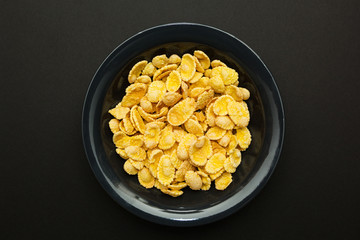 Yellow corn flakes in a black plate on a black background.