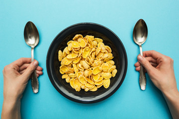 Yellow corn flakes in a black plate on a blue background. Hands with a spoon, eat with both hands. © Andrii Zastrozhnov