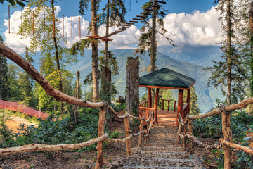 Fototapeta na wymiar Stairway with stony steps leading downward a wooden resting pavilion in adventure rope park in summer forest of Caucasus Mountains. Beautiful scenic landscape with blue sky and clouds