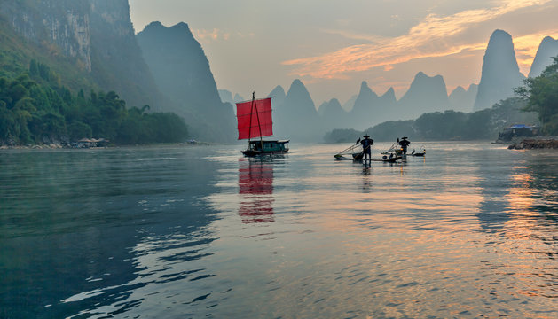 Fisherman stands on traditional bamboo boats at sunrise (boat with a red sail in the background) - The Li River, Xingping, China