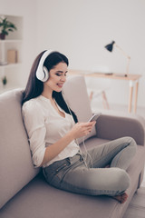 Portrait of beautiful young woman sitting on sofa at home with phone and listen to music with white headphones