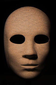 Textured mask with golden wood painted surface, neutral expression on dark background.