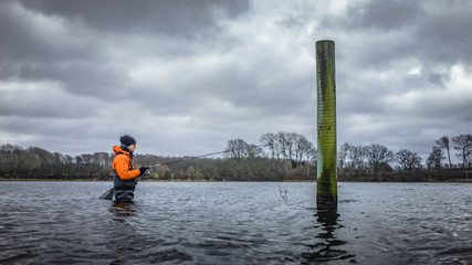 Angler mit Angelrute in Wathose hat Fang im seichten Wasser neben Pfahl vor Küste mit Wald und Bäumen