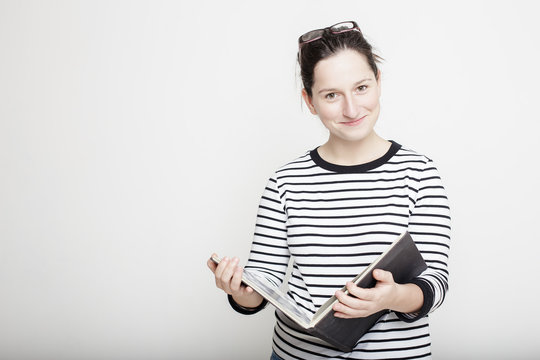 Young attractive smiling woman with glasses on head, standing with a magazine in their hands on a blank gray wall background in a striped sweater