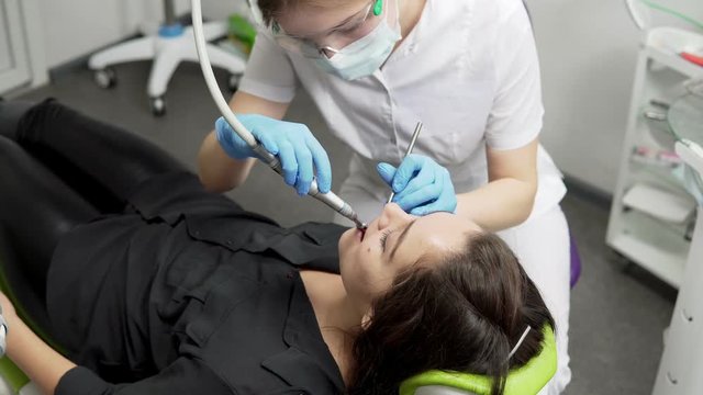 Young female dentist in gloves and mask drilling patient's teeth in clinic