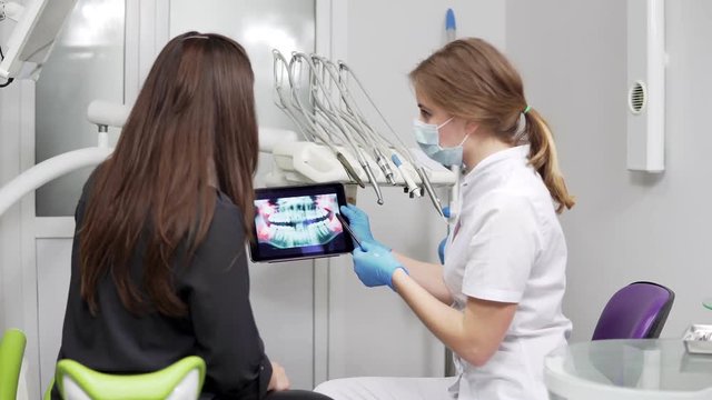 Female doctor dentist is showing x-ray teeth on tablet to young female patient. Dentist is wearing lab coat