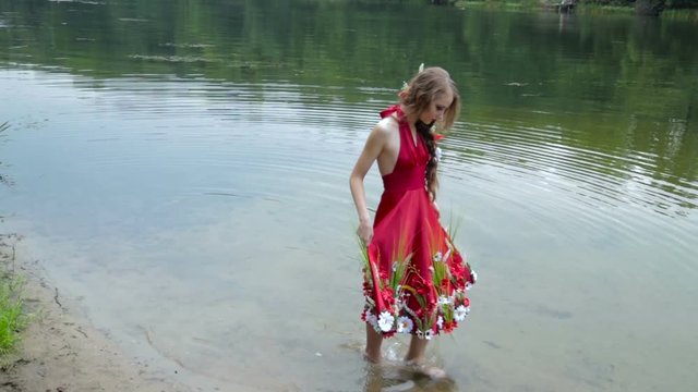 Unusual and mysterious girl with creative make-up and elegant hairstyle with flowers in ethnic red dress walking in water. Fashion, mysterious and nature concept