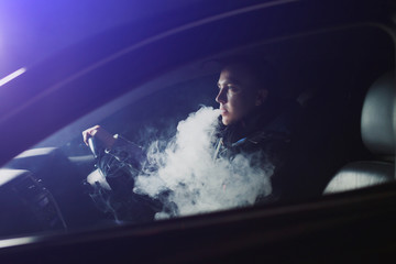 Fototapeta na wymiar View from the side of a young man smoking an e-cigarette as he drives his car on an urban street
