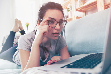 Young surprised woman using laptop while lying on comfortable sofa