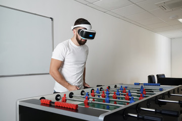 Man in virtual glasses playing at office in table soccer, different technologies concept