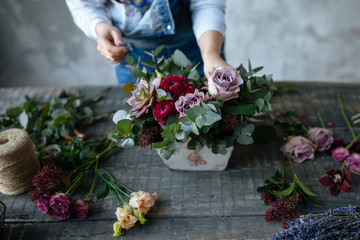 female  in gray blouse and jeans make a bouquet over gray background, putting roses in vase, flowers and vase on wood table, workplace