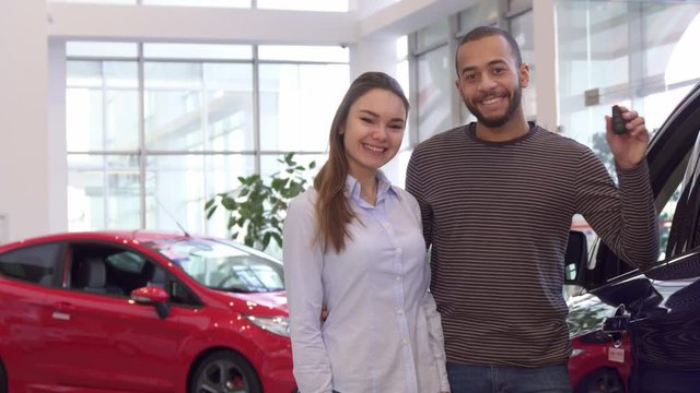 Young interracial couple buying car at the dealership. Handsome african american man embracing his girlfriend against background of red vehicle. Pretty caucasian girl showing her thumb up near her