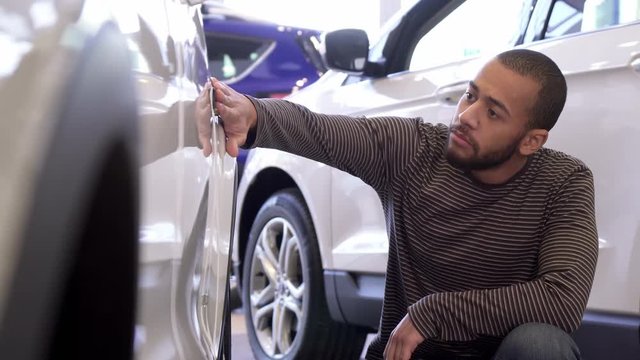 Young black man touching car wing at the dealeship. Low shot of handsome african american guy turning his face to the camera against background of white crossover. Attractive bearded male customer