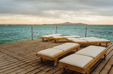Fototapeta na wymiar Pier with chaise longues in the sea in resort. Summer vacation. View at a distant island at sunrise.