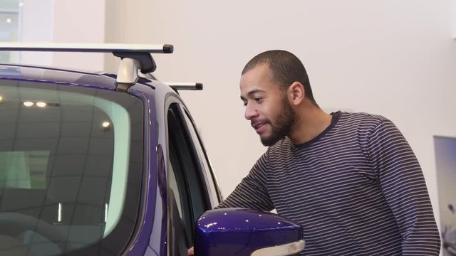 Attractive brunette bearded man standing near the purple crossover at the dealership. Young african american guy examining interior of the car. Handsome male customer demonstrating car key at the