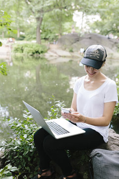 Young woman sitting at lakeside in park using cell phone and laptop