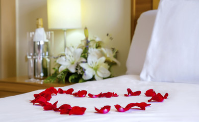 Close-up romantic arrangement with focus on the heart-shaped rose petals. White bedding, flowers, champagne flutes, honeymoon or Valentine's day bedroom set up, or hotel welcome pack for newly weds.