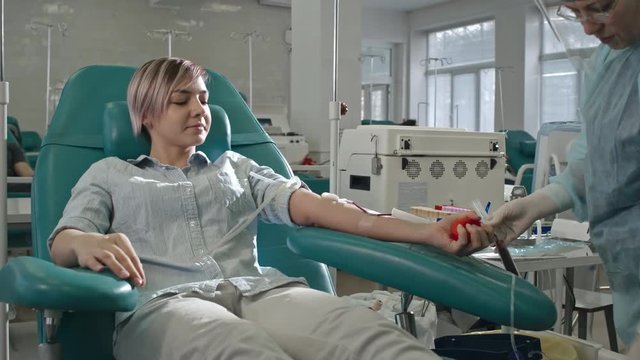 Pretty young woman sitting on medical chair while nurse taking her venous blood in test tube