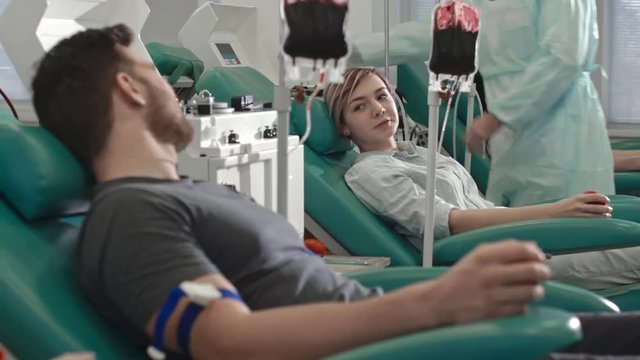 Tracking shot of young woman and man sitting in medical chairs and chatting while donating blood in hospital 