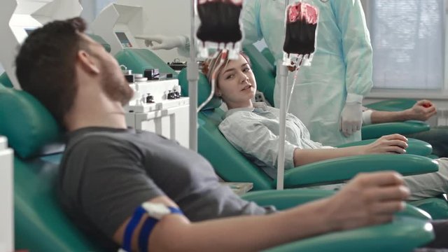 Tracking shot of young woman and man on medical chairs donating blood in hospital. They looking at each other and chatting
