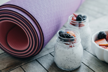 Sport and healthy concept. Pink yoga mat and Pudding with chia seeds, yogurt and fresh fruits: Strawberries, blueberries and blackberries in glass jars on wooden background