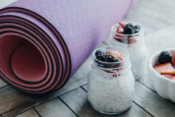 Schilderijen op glas Sport and healthy concept. Pink yoga mat and Pudding with chia seeds, yogurt and fresh fruits: Strawberries, blueberries and blackberries in glass jars on wooden background © Boyarkina Marina