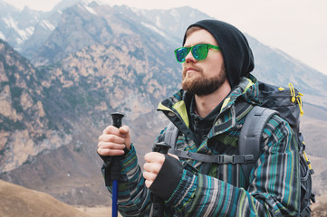 A guy with a beard and wearing sunglasses in a membrane jacket, hat, with a backpack and sticks for Nordic walking, a traveler standing and looking at the mountains