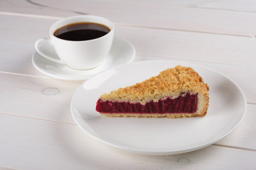 A cup of black coffee and a piece of tasty berry pie on a plate stand on a white wooden table