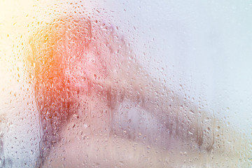 Beautiful young woman washes in the shower behind the glass in water droplets