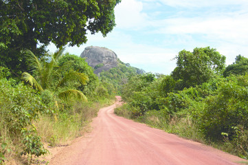 Dusty road going through green colored bushes leading to a big black rock at midday and nobody around on the island Belitung, Indonesia.